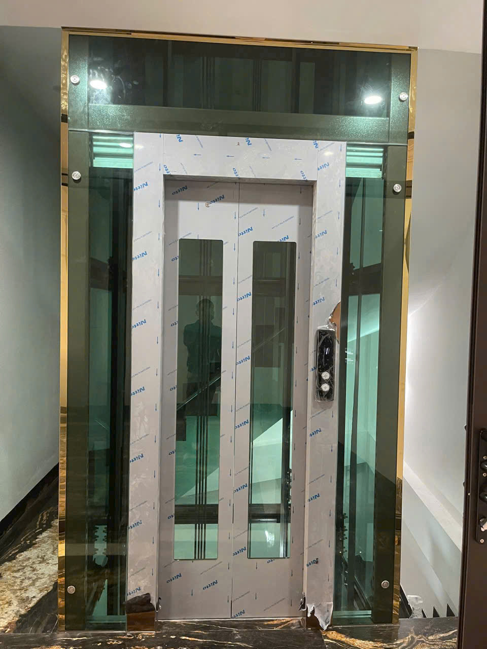 Thang máy gia đình vách kính khung viền mạ vàng đang lắp đặt trong nhà